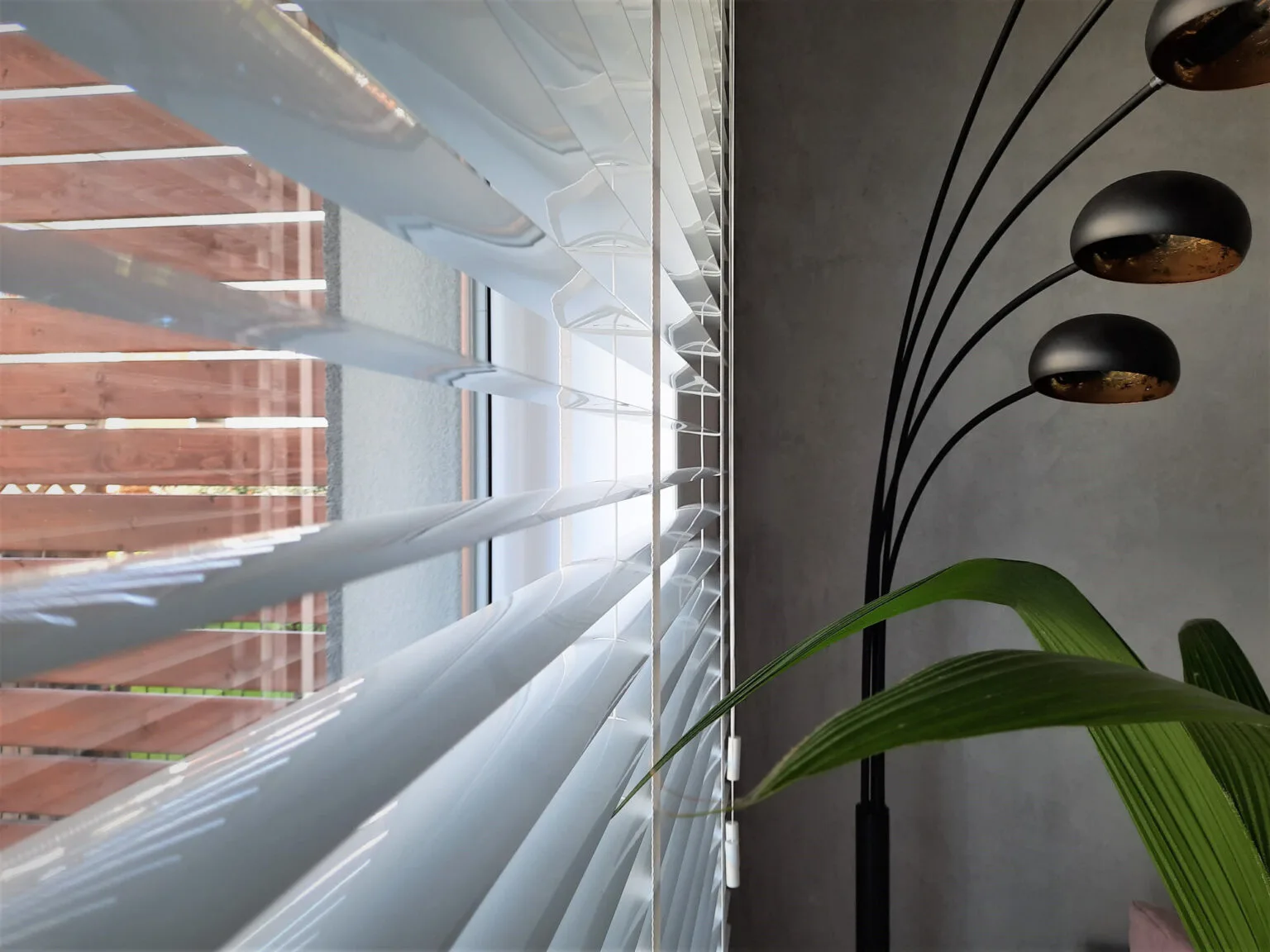 Wyczyszczone żaluzje aluminiowe białe z drabinką taśmową koloru białego. Zbliżenie na lamele aluminiowe żaluzji wewnętrznej. Z prawej strony widoczna na tle szarej betonowej ściany nowoczesna czarna 5-ramienna lampa oraz roślina doniczkowa z zielonymi liśćmi. Zdjęcie zostało prawdopodobnie wykonane w jednej z miejscowości, w obrębie których Perfecta Home montuje swoje produkty np. Wrocław, Oława, Brzeg Dolny, Trzebnica, Kąty Wrocławskie, Kobierzyce, Syców, Oleśnica, Legnica, Środa Śląska, Oborniki Śląskie, Jelcz Laskowice